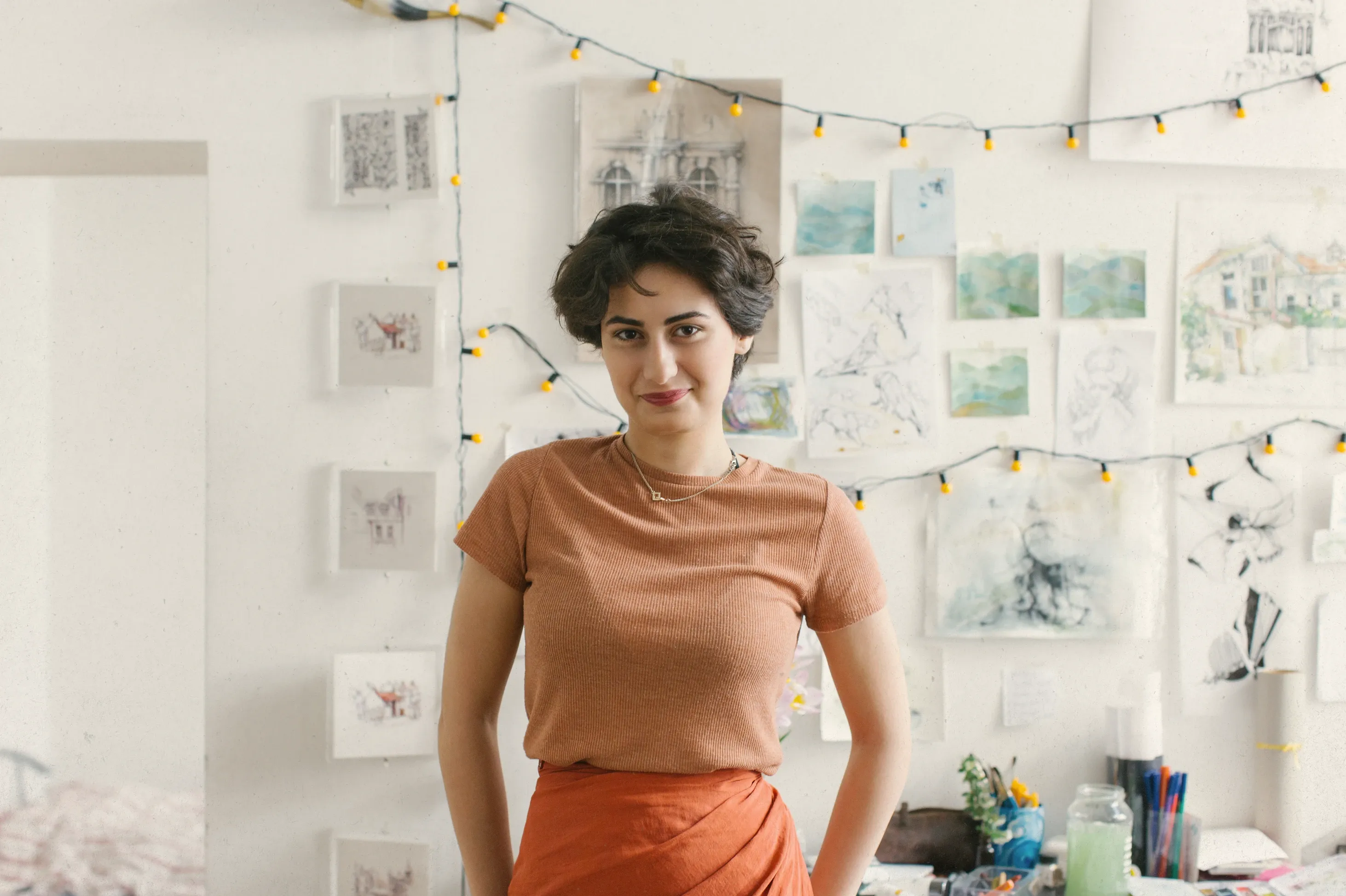 An artist standing in her studio, sketches pinned to the wall behind her